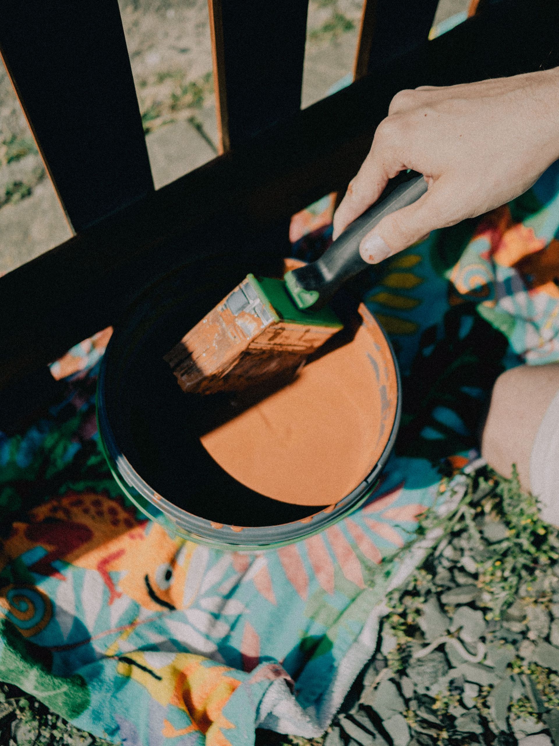 Close-up of a person painting a wooden fence with a brush and orange paint outdoors on a sunny day.